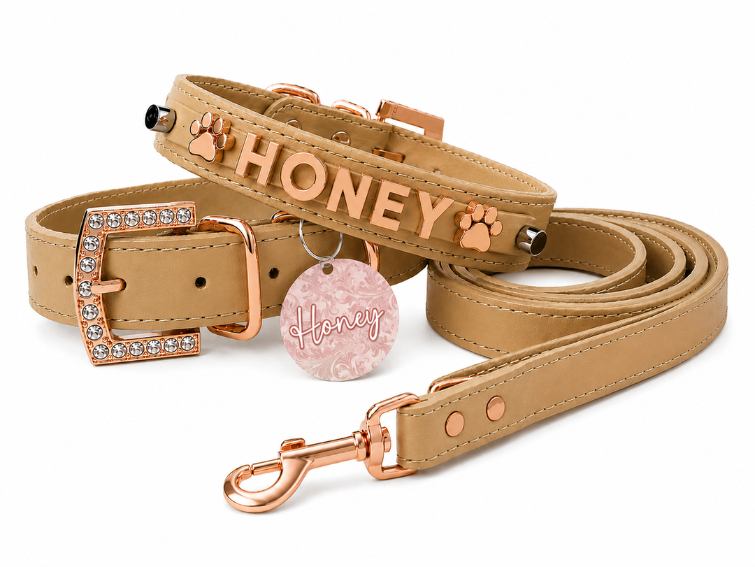 A tan faux-leather pet collar and matching leash set with rose gold hardware. The collar is personalized with the name 'HONEY' in raised rose gold letters, flanked by two paw prints, and features a matching round pink marbled tag and a rhinestone-embellished buckle.